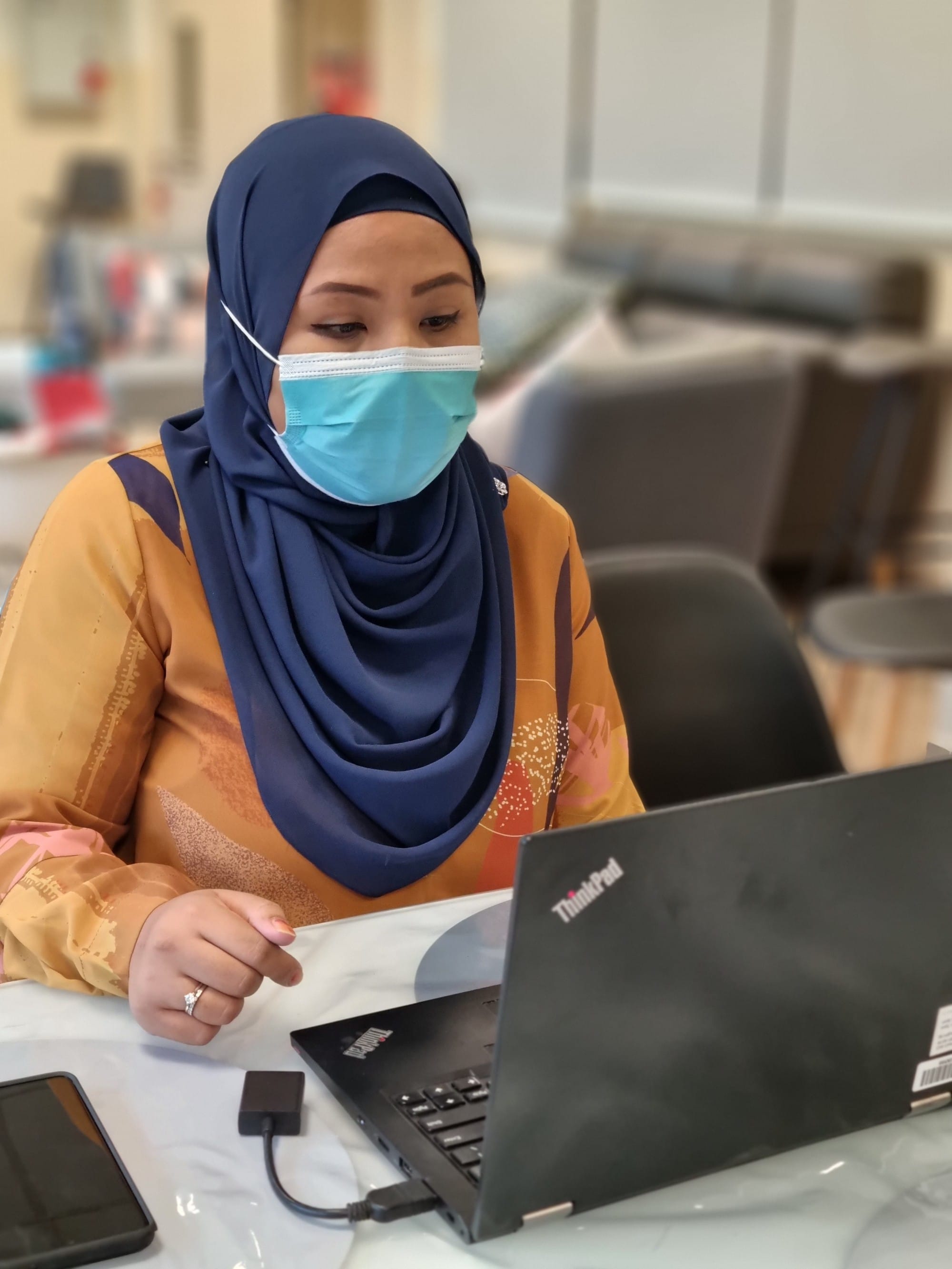 Woman wearing a navy hijab and blue face mask sits at a table working on a ThinkPad laptop.